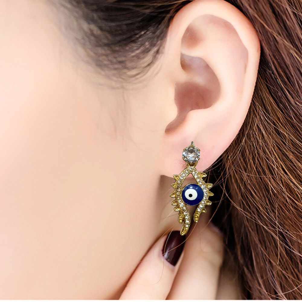 Close-up of gold-tone evil eye earrings with sparkling crystal embellishments on a red background
